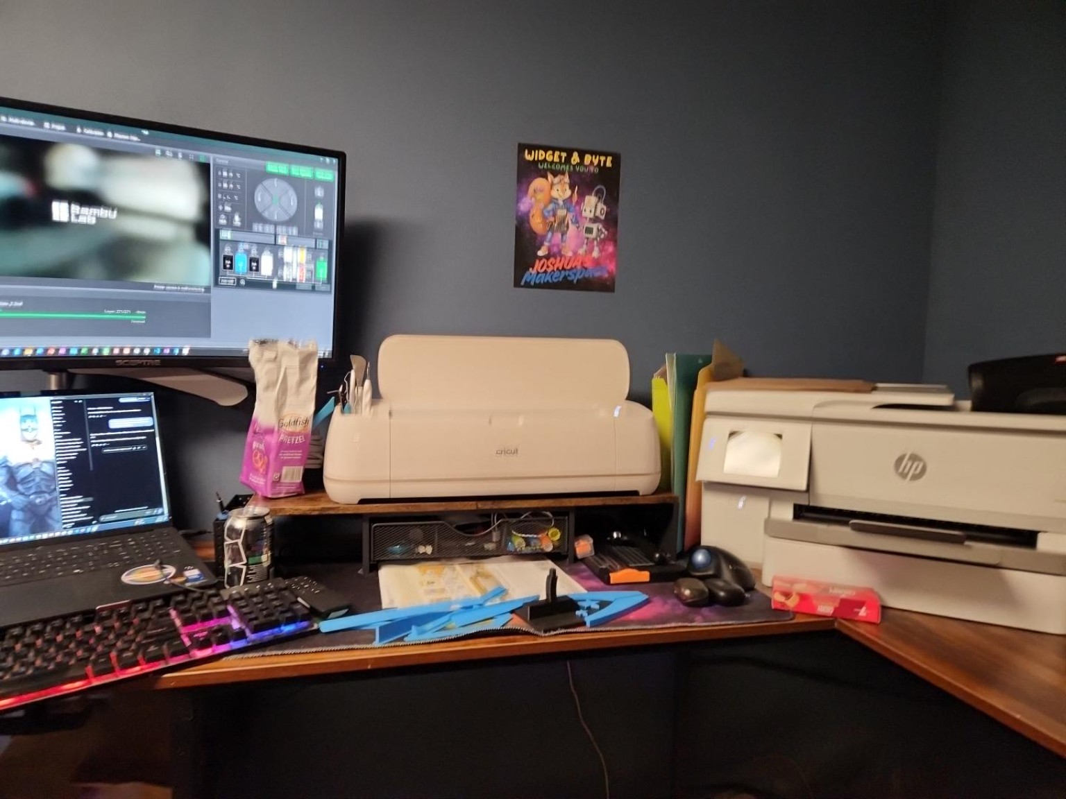 My design, cutting, and printing workstation with a pale Cricut machine, an HP wide-format printer, monitors, a laptop, keyboard, tools, and a Widget and Byte Makerspace print on the wall.